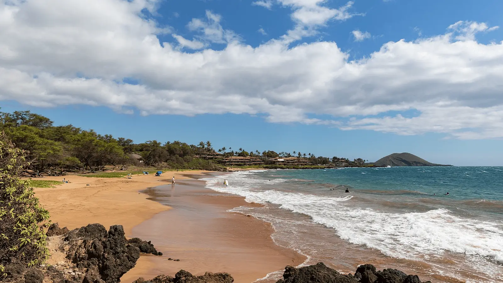 A scenic beach with golden sand, gentle waves, and rocky outcrops in the foreground. People are enjoying the water and walking along the shore. In the background, there are lush green trees and resort buildings beneath a blue sky with some clouds—perfect for snorkeling on Maui.