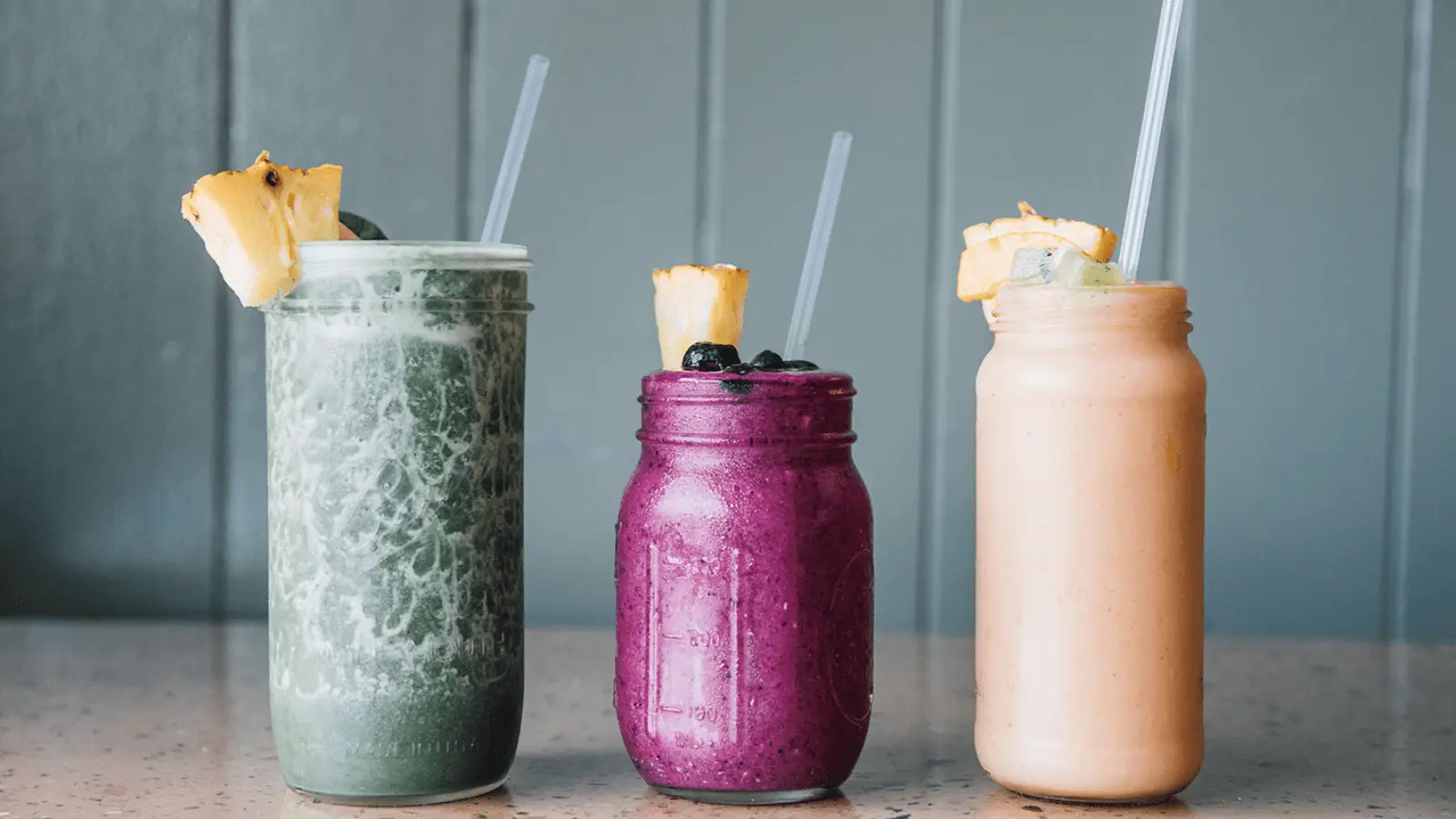 Three mason jars filled with different smoothies are lined up on a speckled countertop, making for the best breakfast. The smoothies, from left to right, are green, purple, and orange. Each jar is topped with a slice of pineapple and has a straw—just like you'd find on Kauai's South Shore.