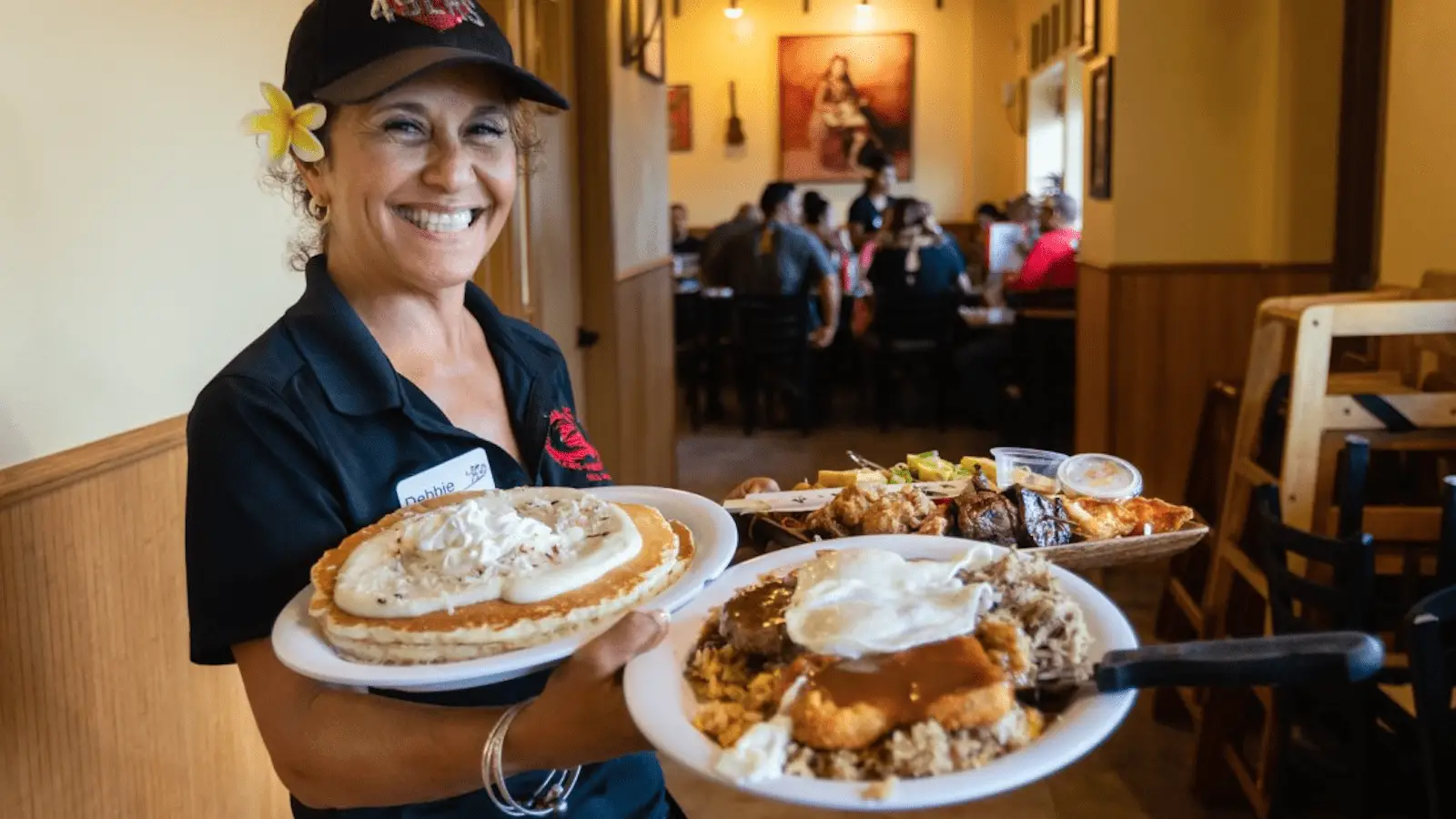 A smiling waitress with a flower in her hair holds three plates of food inside a restaurant. One plate has pancakes with whipped cream; another has a dish with sauce and rice, and the third contains various items, including grilled meat with accompaniments. Diners enjoying the best breakfast on the Big Island are seated in the background.