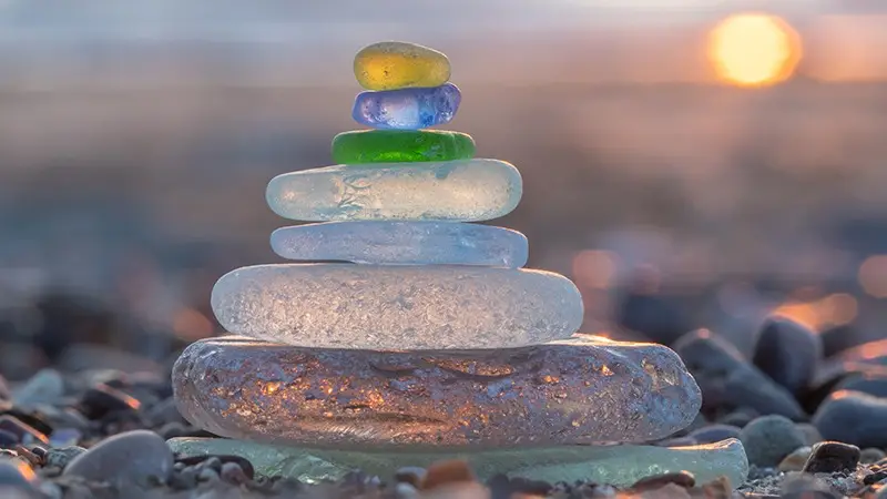 A stack of colorful glass pebbles of varying sizes balanced on a rocky beach with a soft-focus background captures the warm glow of the setting sun. The pebbles form a gradient from clear at the bottom to shades of blue, green, and yellow towards the top—one of the best things to do on Monterey Peninsula in November.