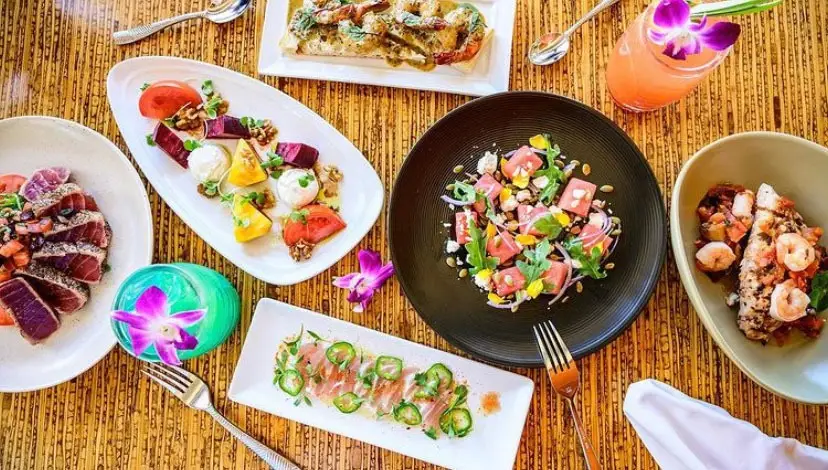 A colorful assortment of gourmet dishes is arranged on a wooden table. Plates include seared tuna, caprese salad, a seafood dish with shrimp, a mixed poke bowl, jalapeño-covered sashimi, and a slice of dessert with an orchid flower garnish. A drink with an orchid completes the setting—perfect for the best breakfast in Kaanapali.