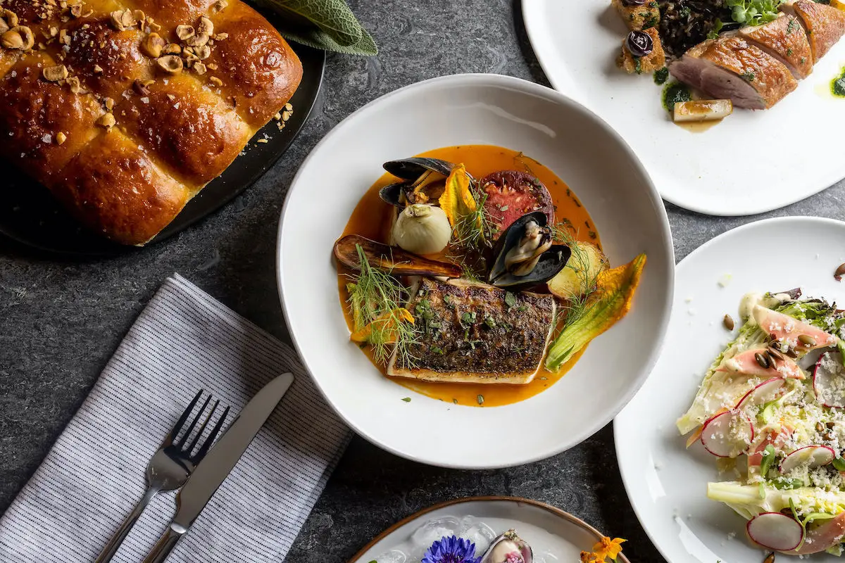 An overhead view of a table setting featuring various dishes suggests one of the best dinner spots in Sonoma County. The main dish in the center appears to be grilled fish garnished with vegetables and sauce. Surrounding it are plates with bread, a meat dish, and a salad, along with a striped napkin, knife, and fork.