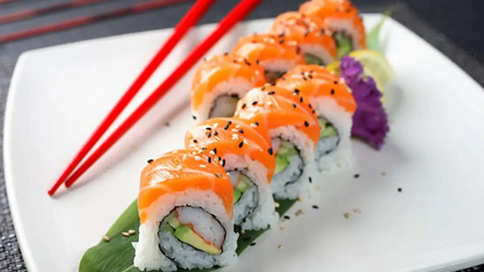 A neatly arranged sushi roll topped with slices of raw salmon, garnished with black sesame seeds. The roll is placed on a green leaf on a white plate, accompanied by red chopsticks. A small amount of purple decoration and a lime wedge are on the side, showcasing why it’s the best sushi Kauai offers.