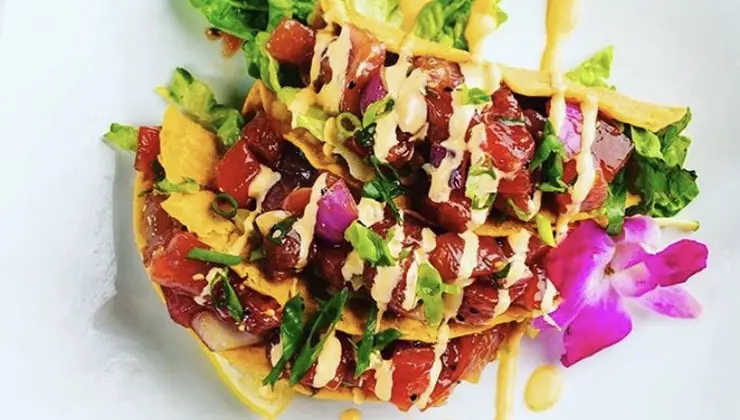 A vibrant plate of ahi tuna nachos garnished with green onions, drizzled with a creamy sauce, and served with fresh lettuce and edible purple flowers. Reminiscent of the best poke from Waikiki Oahu, the tuna is cut into small, bite-sized pieces atop crispy nacho chips.