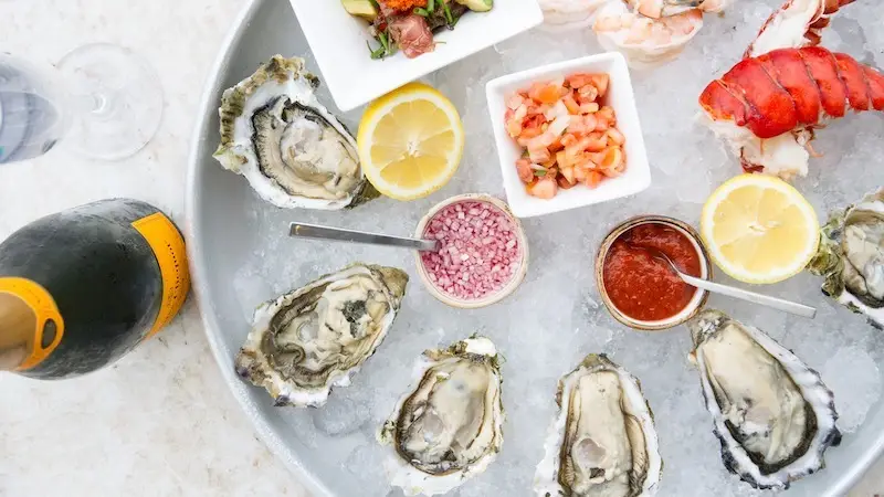 A platter of seafood featuring six oysters, a lobster tail, and shrimp, garnished with lemon halves and small bowls of sauces. Two lemons, diced tomatoes, and a small cup of red onions are arranged on the plate. A partially visible bottle of champagne is on the left—the best happy hour Waikiki has to offer.