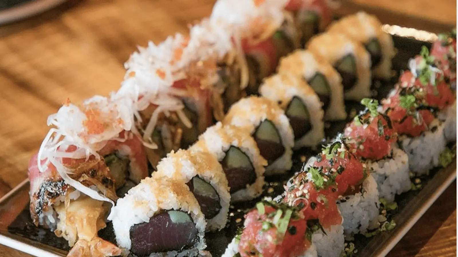 A rectangular black platter holding an assortment of sushi rolls on a wooden table. The rolls are garnished with ingredients including tuna, cucumber, crab, and various toppings such as tempura flakes, shredded crab, and chopped green onions. Truly the best sushi Kauai has to offer.