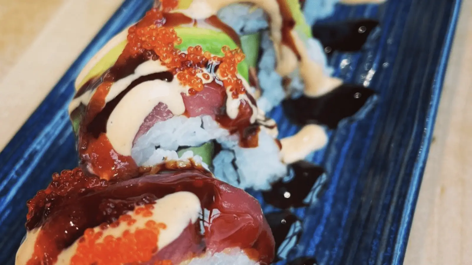 A close-up of the best sushi on the Big Island, beautifully arranged on a dark blue rectangular plate. The rolls are topped with various ingredients including raw fish slices, avocado, orange fish roe, and drizzles of creamy sauce and dark sauce, creating a colorful and appetizing presentation.
