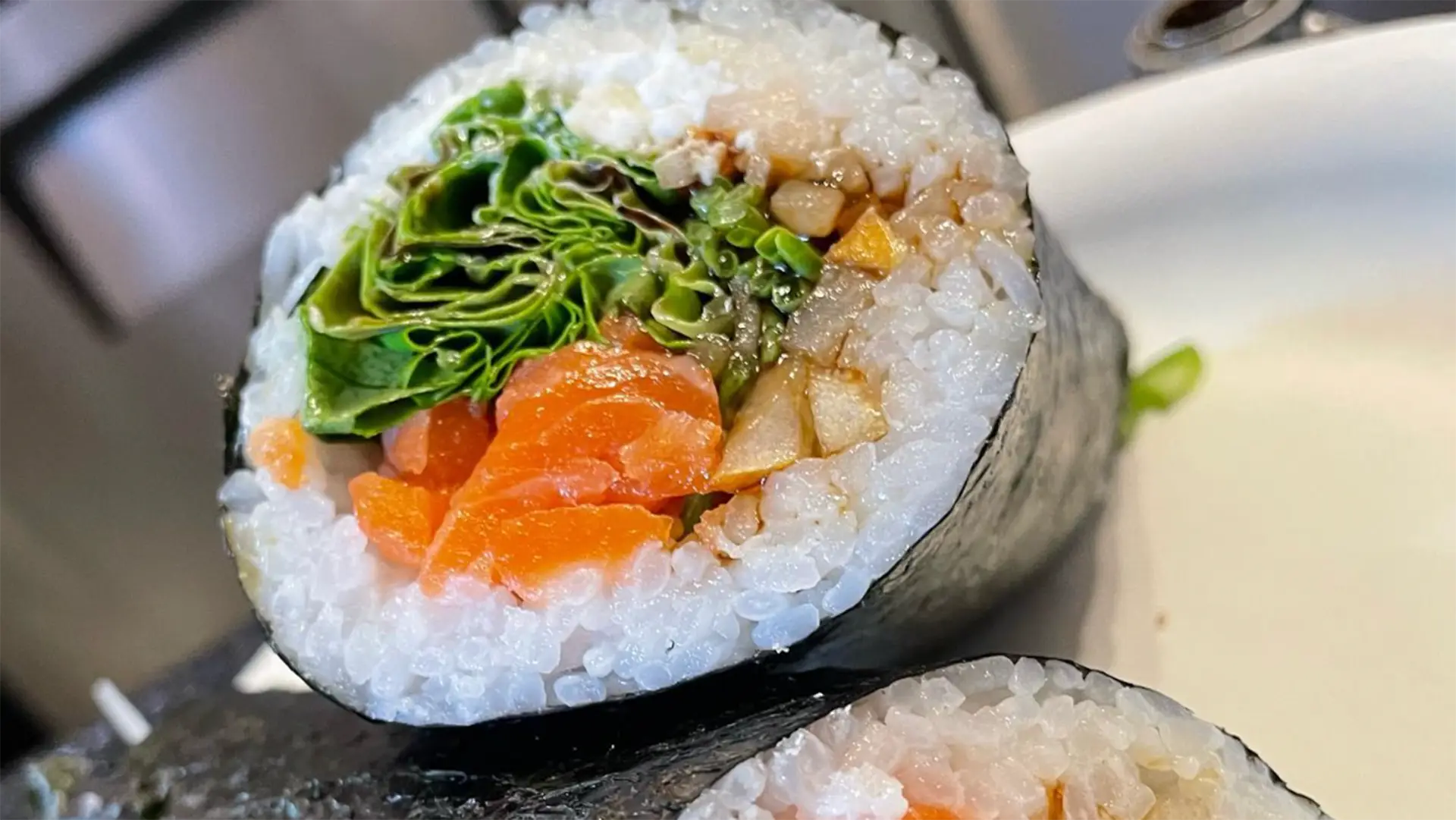 Close-up of two sushi rolls. The roll in the foreground is filled with white rice, green leafy vegetables, orange fish, and brown sauce, wrapped in seaweed. The second roll is partially visible in the background with similar fillings. These tempting rolls rest on a white plate—possibly some of the best sushi Kauai offers.