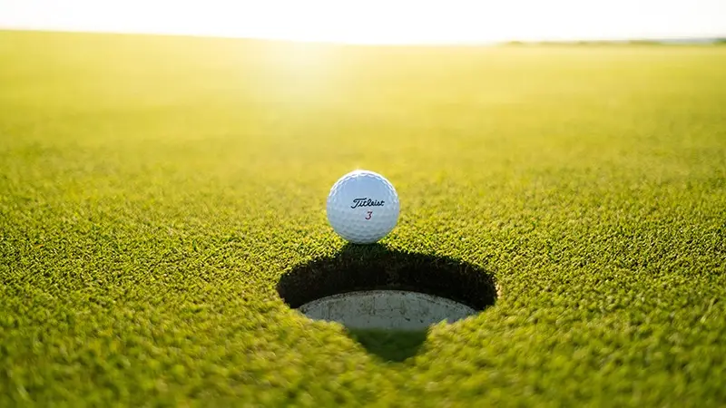 A close-up shot of a white golf ball resting at the edge of a hole on a well-maintained green golf course. The sun is shining brightly, casting a soft light and creating a shadow of the ball over the hole, emphasizing the precision required for the putt. Among the best things to do on Monterey Peninsula in November is enjoying such pristine golfing conditions.