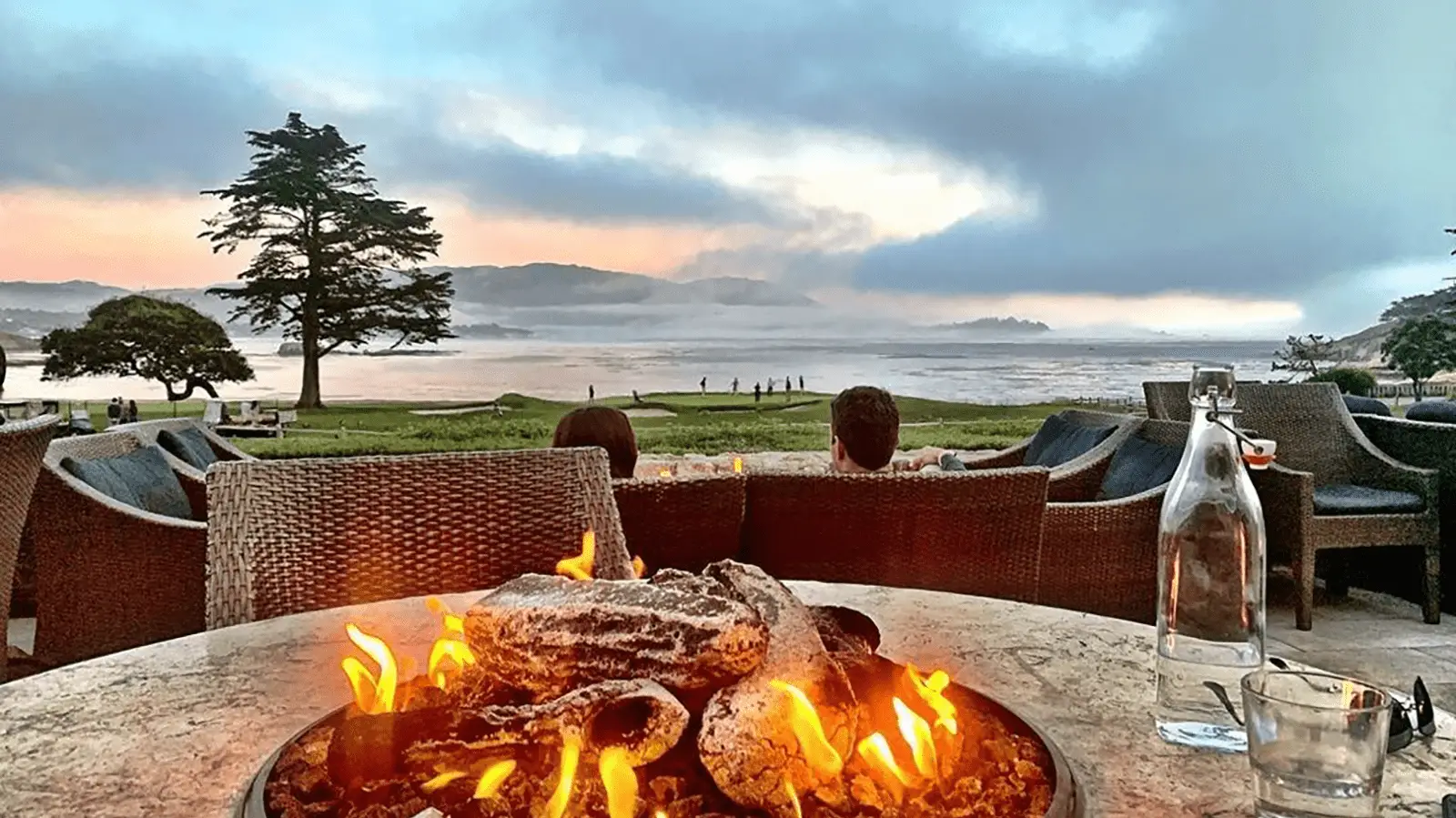 A cozy fire pit burns brightly in the foreground, surrounded by outdoor wicker seating. Beyond the fire, people can be seen standing and walking near a tranquil body of water, with a dramatic, cloudy sky and distant hills completing what could be the best restaurant view on Monterey Peninsula.