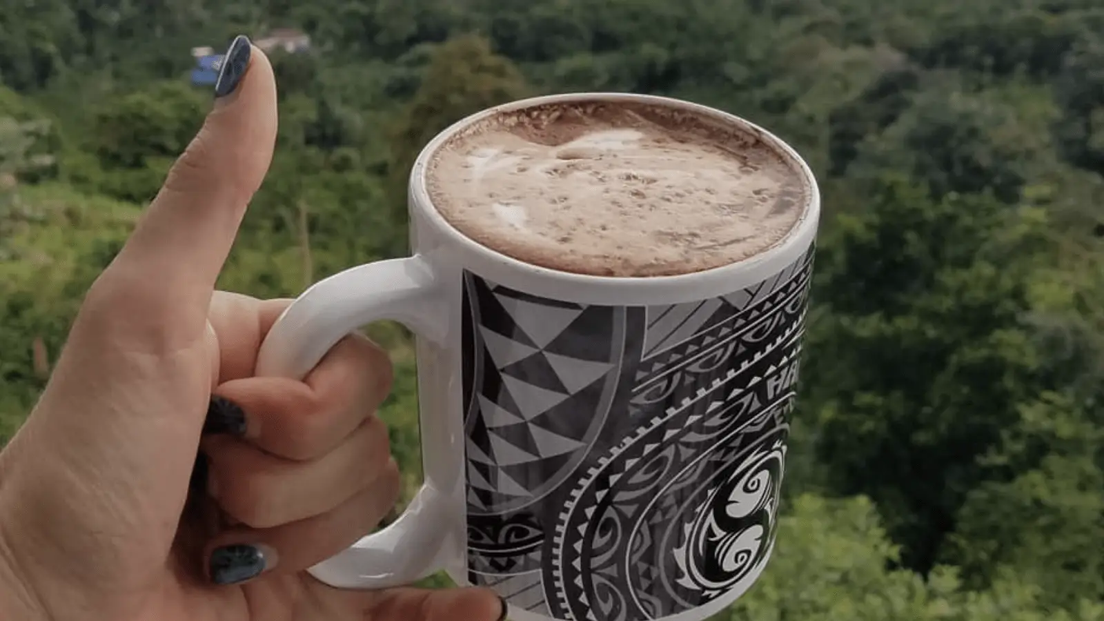 A hand with painted nails holds a white mug with intricate black patterns, filled with frothy hot chocolate. In the background, a lush, green forest, slightly out of focus, creates a serene and natural setting—truly reminiscent of the best breakfast on the Big Island.