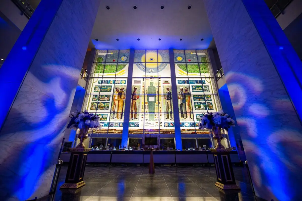 A grand hall with tall, colorful stained-glass windows depicting various figures and scenes illuminated by blue lighting. The floor reflects the vibrant lights. Ornate flower arrangements on pedestals flank the central aisle, leading to an area with display counters—a highlight in "Best Things to Do in San Francisco May.
