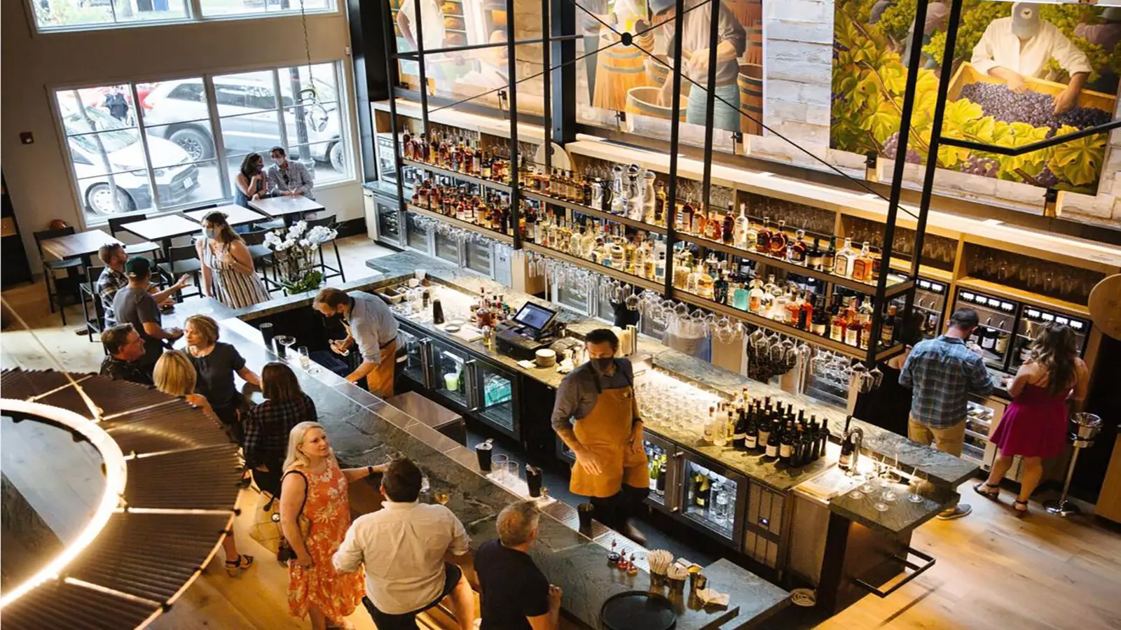 A lively bar scene in one of the best dinner spots in Sonoma County, a modern restaurant with high ceilings. Patrons chat at the bar and tables near large windows. A bartender in an apron serves drinks. Colorful murals adorn the walls above the bar, and natural light fills the space.