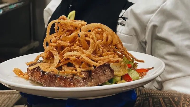 A person in a chef's coat holds a white plate with a steak topped with a generous amount of crispy fried onions. The steak is served on a bed of mixed vegetables including sliced cucumbers, red bell peppers, and other greens, perfect for enjoying during the best happy hour Oahu has to offer.