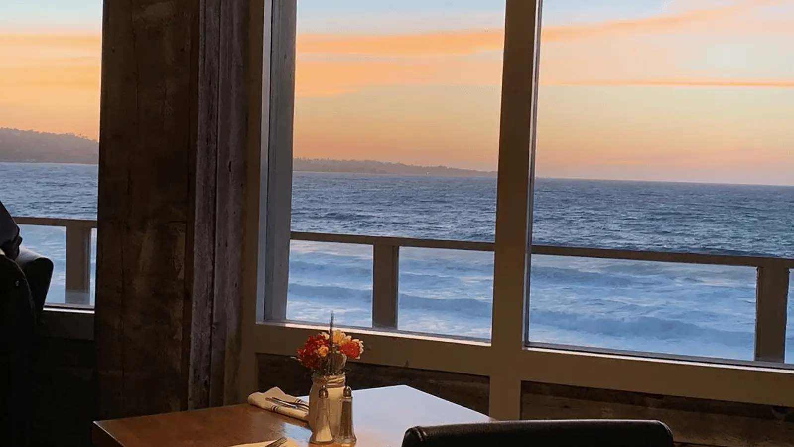 A wooden table with a small vase of flowers is set next to a large window showcasing a beautiful ocean view at sunset—the best restaurant view on the Monterey Peninsula. The sky glows with soft orange and pink hues, and gentle waves can be seen in the distance. A chair is partially visible beside the table.