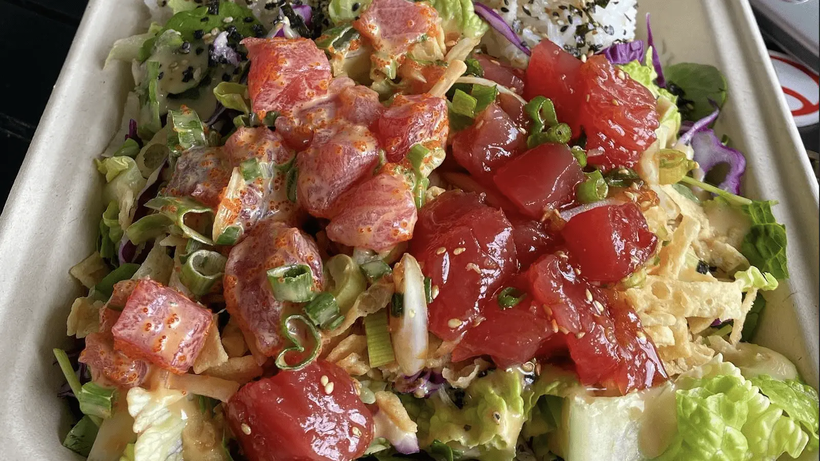 A poke bowl brimming with an assortment of fresh ingredients, including chunks of raw tuna, chopped green onions, shredded cabbage, and lettuce. Garnished with sesame seeds, avocado, and drizzled with a creamy spicy sauce—it's the Best Poke Maui can offer.