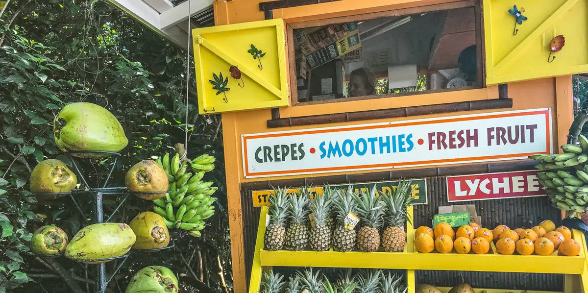 A colorful fruit stand set against lush greenery displays an assortment of tropical fruits, including coconuts, bananas, pineapples, and mangoes. A sign on the stand reads, "Top Foodie Tours Oahu: Crepes, Smoothies, Fresh Fruit, Lychee" in vibrant letters.