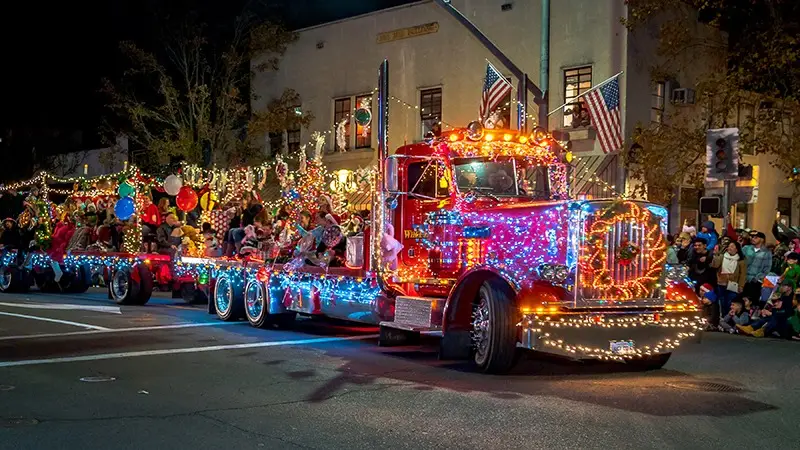 A decorated truck adorned with colorful lights, balloons, and festive decorations drives down a street during a nighttime parade. The flatbed is filled with people in costumes, and American flags are displayed prominently. Spectators line the street, enjoying one of December's best events in Napa Wine Country.