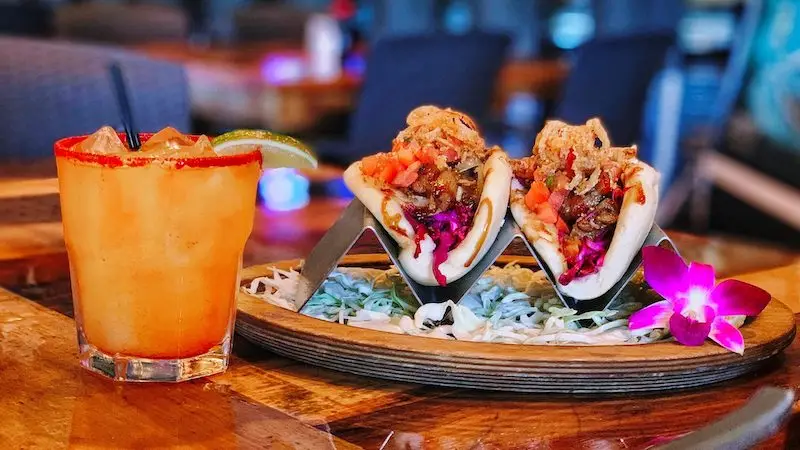 A vibrant plate of two tacos filled with colorful ingredients is displayed on a wooden platter beside an orange cocktail garnished with a lime wedge. An orchid flower is placed next to the tacos on the platter, adding a touch of elegance—perfect for the best happy hour on the Big Island.