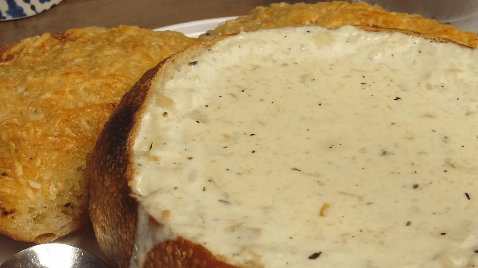 A close-up of a bread bowl filled with creamy, white clam chowder, garnished with herbs. Behind it, two toasted bread slices are visible. A spoon is partially seen at the lower left corner. The texture of the soup and bread is detailed and appetizing—a nod to the best clam chowder on the Monterey Peninsula.