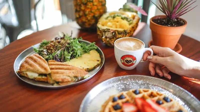 A hand holding a cup of coffee is placed next to plates of food on a wooden table. The plates include a fresh salad with sandwiches, a pineapple bowl filled with pineapple chunks, and a waffle topped with blueberries and strawberries—a scene perfect for the Best Breakfast Waikiki has to offer. A potted plant is in the background.