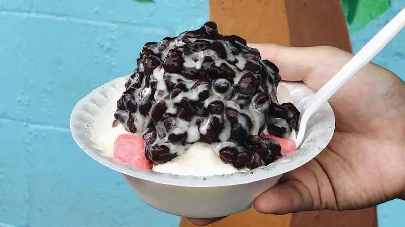 A person holds the best shave ice in Waikiki in a white plastic bowl, filled with shaved ice topped with sweet red beans and drizzled with condensed milk. Pink mochi pieces rest at the base, and a white plastic spoon is placed inside. The backdrop features a blue wall.