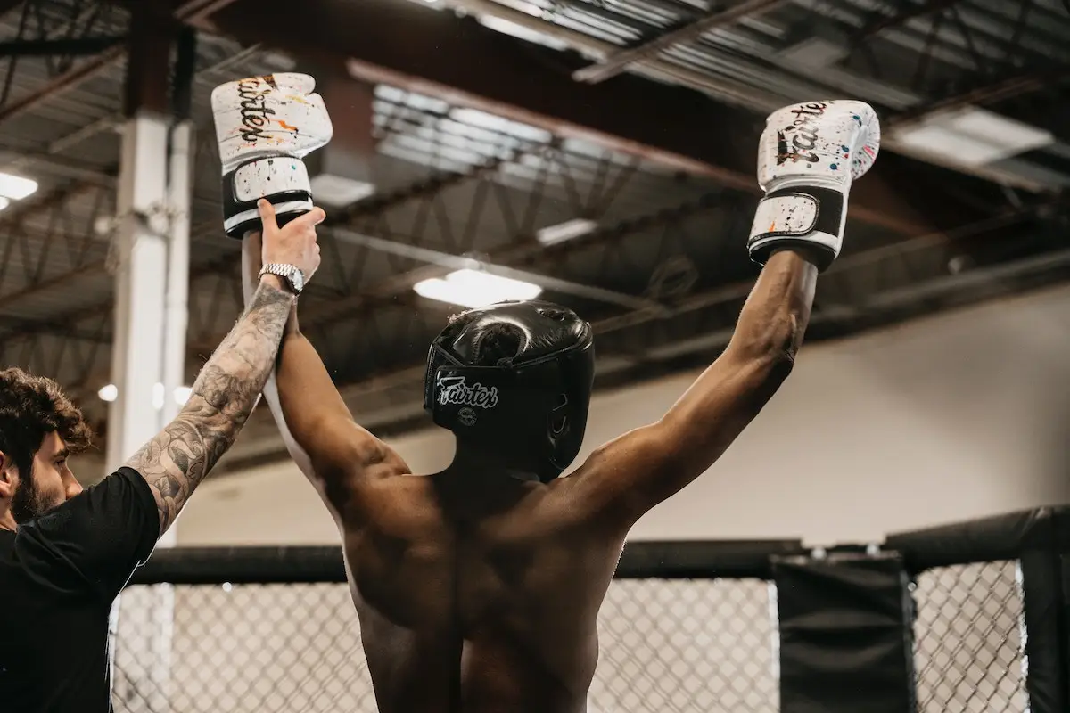 A boxer wearing a headguard and gloves raises his arms in victory inside a cage-like ring. An individual with tattooed arms stands beside him, also raising one of the boxer's arms. It’s like one of the best events to do in November on the Monterey Peninsula, against an industrial-style ceiling and lighting backdrop.