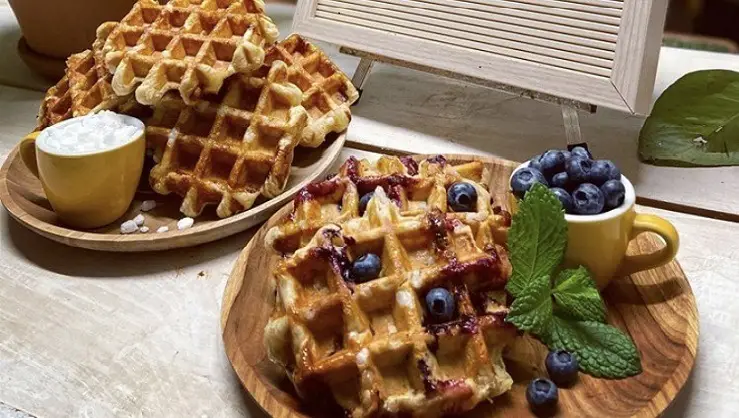 A wooden table displays a wooden plate with a waffle topped with blueberries, a small mint garnish, and a yellow cup filled with blueberries. In the background, more waffles are stacked on a plate beside a small yellow bowl with white cream. A blank letter board stands upright showcasing the best breakfast North Shore.