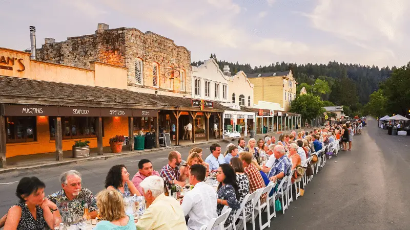 A long table is set up on a street with numerous people sitting and enjoying a meal together in an outdoor communal dining setup. The background features historic buildings and storefronts under a partly cloudy sky, giving the scene a lively and rustic atmosphere—one of the best things to do in Napa Valley this September.