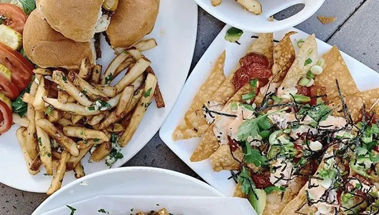 A table with various dishes, perfect for the best happy hour Waikiki offers, includes a plate of burgers and fries on the left, a plate of nachos topped with sliced green onions, seaweed, and other garnishes on the right, and the edge of another plate containing fries partially visible at the bottom.