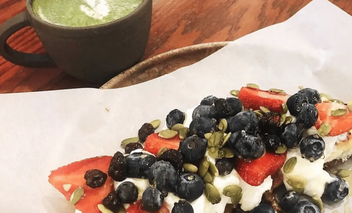 A slice of toast topped with strawberries, blueberries, pumpkin seeds, and a drizzle of yogurt is placed on a brown parchment paper. Beside the toast, there's a cup of matcha latte with frothy milk in a dark ceramic mug on a wooden surface.