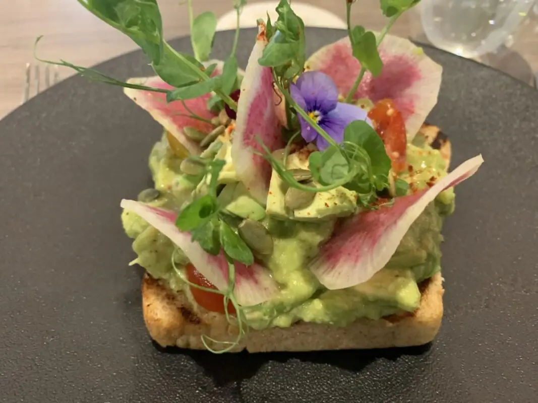 A gourmet dish featuring avocado toast on a dark plate. The toast is topped with mashed avocado, microgreens, watermelon radish slices, pumpkin seeds, and an edible purple flower, creating a colorful and visually appealing presentation.