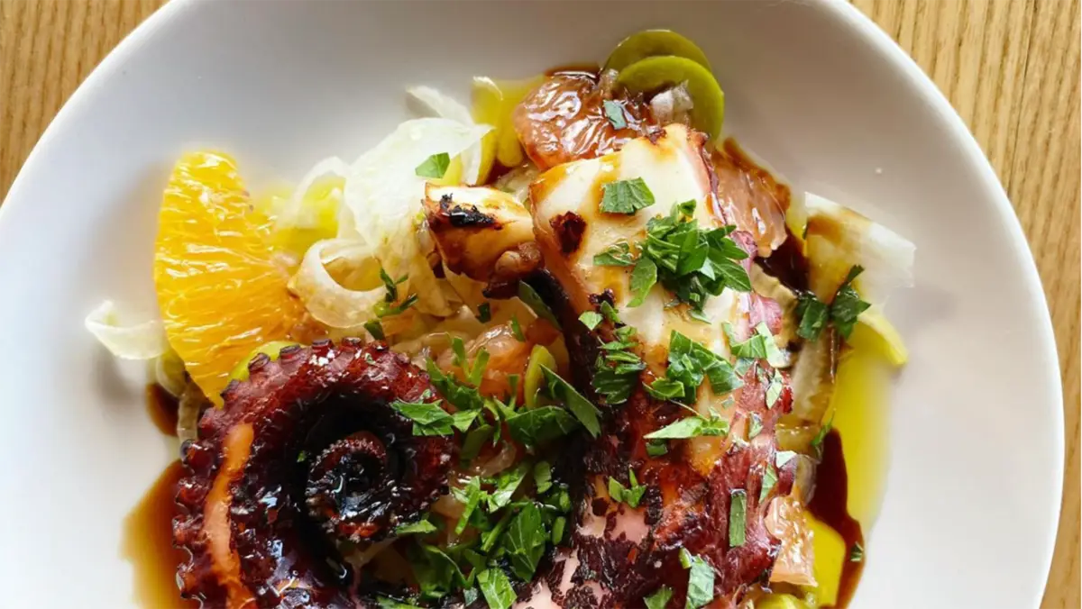 A gourmet dish featuring grilled octopus garnished with chopped fresh herbs on top, perfect for the best dinner in Princeville. The octopus tentacles are served alongside thin slices of bright orange and pale yellow vegetables, all plated on a white dish. The presentation is vibrant and appetizing.