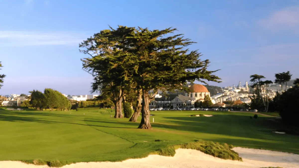 A scenic view of one of the best golf courses San Francisco boasts, with green fairways, sand bunkers, and tall trees. In the background, there's a large domed building and an urban skyline under a clear blue sky. This is just one of the many things to do in the Bay Area for golfing enthusiasts.