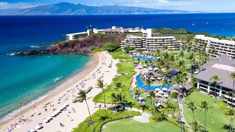 Aerial view of a tropical beach resort with a long sandy beach lined with umbrellas and lounge chairs. Green lawns and palm trees surround the resort buildings, which include several swimming pools. The deep blue ocean stretches to the horizon with mountains in the background, reminiscent of the best beaches for walks on Maui.