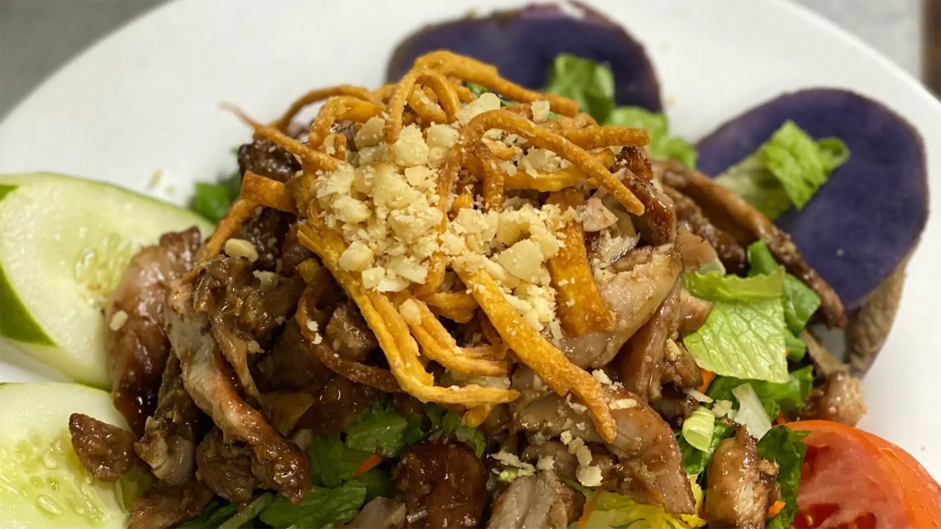 A close-up of a salad topped with a generous serving of grilled chicken strips, crispy fried noodles, and crushed peanuts. The salad includes fresh greens, tomato slices, cucumber slices, and purple potato slices, all arranged on a white plate. Truly the best Hawaiian food on the Big Island.