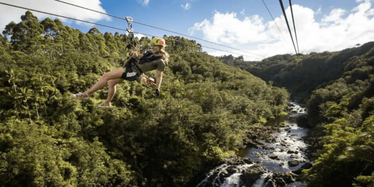 A person wearing a helmet and harness is zip-lining over a lush green forest on one of the Top Big Island Zip Line Tours. Below, a river flows through the forest with rocks and small waterfalls, and the sky above is mostly clear with a few clouds. The scene looks adventurous and serene.
