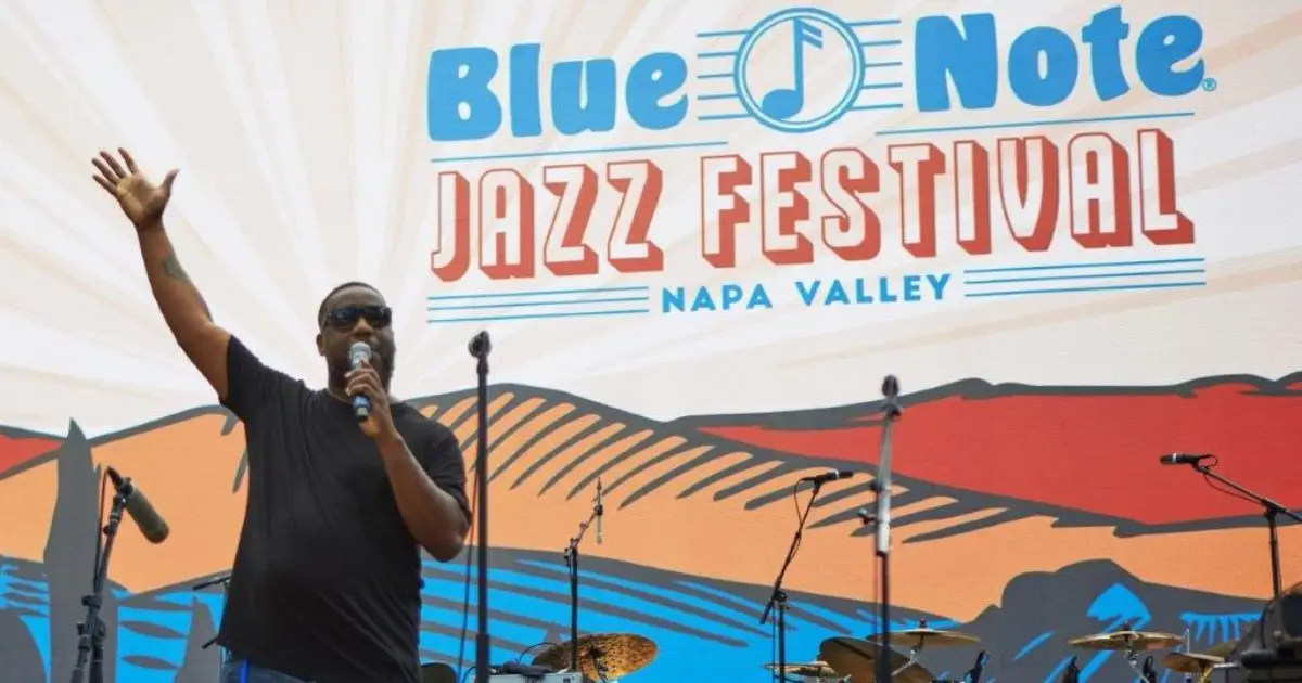 A performer wearing sunglasses and a black shirt holds a microphone and raises one arm on stage at the Blue Note Jazz Festival in Napa Valley, one of the best things to do in July. The festival's colorful backdrop features the event name and stylized images of mountains and sky, with musical equipment on stage.