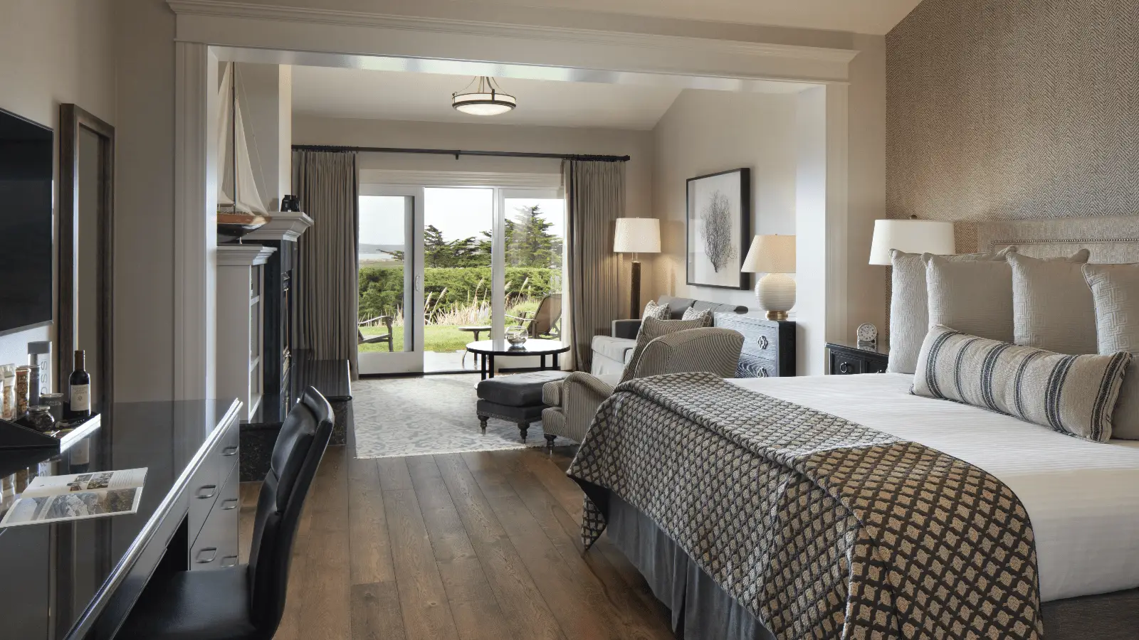 A modern, well-lit hotel room at Bodega Bay Lodge features a large bed with patterned bedding, a cozy seating area with a grey sofa and armchairs, a desk, a flat-screen TV, and a balcony with views of green landscaping and trees. The decor includes neutral tones and elegant furnishings.