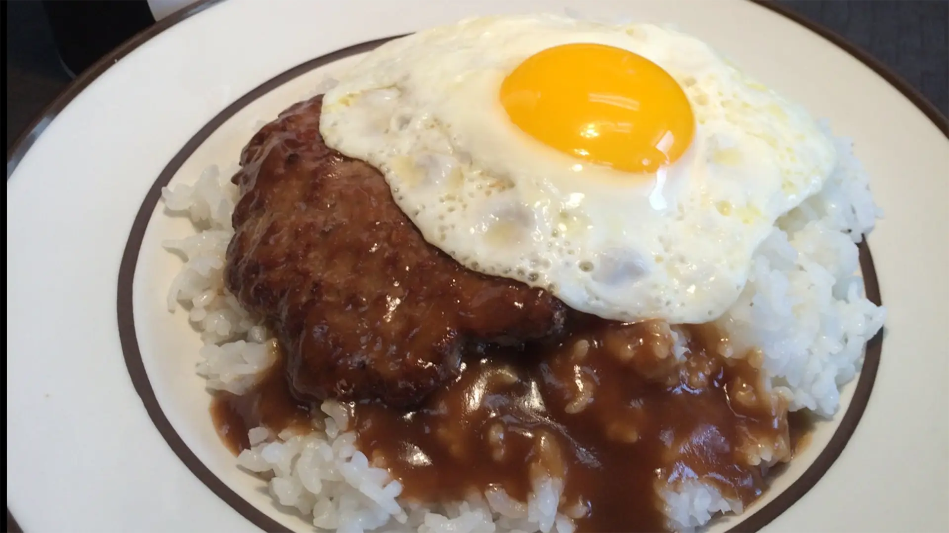 A plate of Loco Moco, featuring a beef patty on a bed of white rice, topped with a fried egg and gravy. The yolk of the egg is bright yellow and perfectly cooked. The brown gravy surrounds the beef patty, creating a savory presentation on a white plate—truly some of the best Hawaiian food on the Big Island.