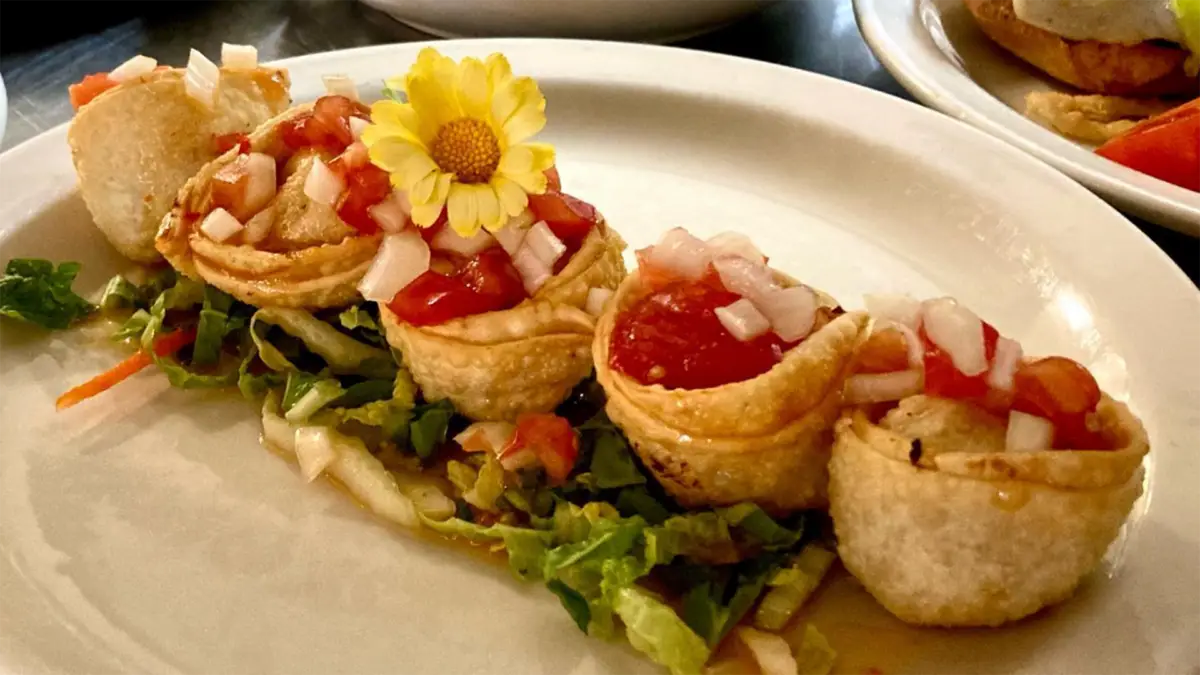 Four beautifully presented appetizers are arranged in a line on a white plate. Each is topped with diced tomatoes and onions, sitting on a bed of shredded lettuce and carrots, making it the best lunch on the Kohala Coast. A small yellow flower is placed decoratively beside them.