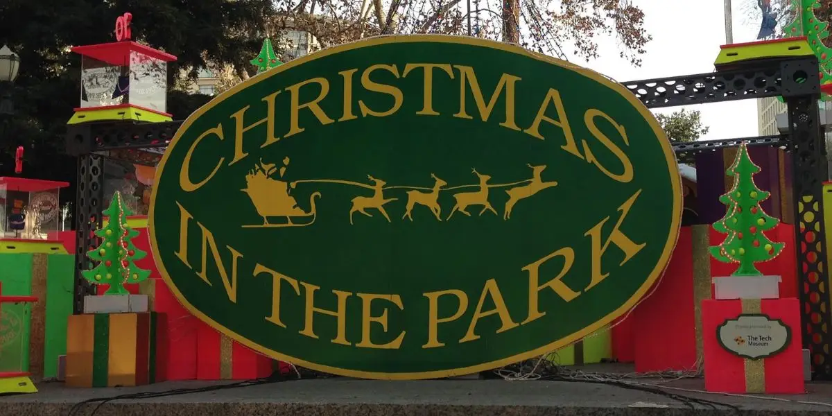 A large green oval sign with the words "CHRISTMAS IN THE PARK" in yellow letters features a silhouette of Santa's sleigh pulled by reindeer. Surrounded by colorful holiday decorations, including small Christmas trees and presents, this event is one of the best things to do in the Bay Area this December.
