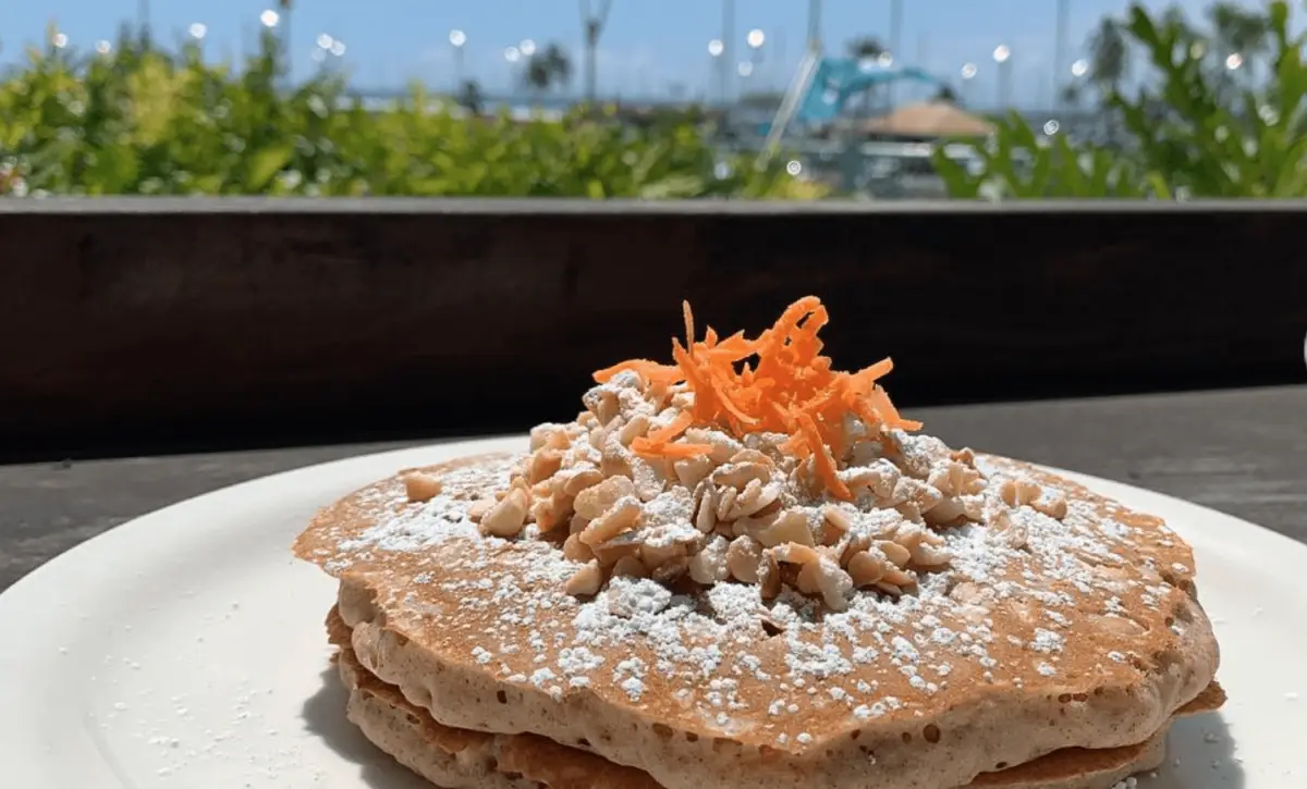 A plate of pancakes topped with grated carrots, chopped walnuts, and powdered sugar is set on a table outdoors. The background shows a sunny, scenic view with greenery, palm trees, and a marina with boats docked in the distance - truly the best breakfast Waikiki has to offer.