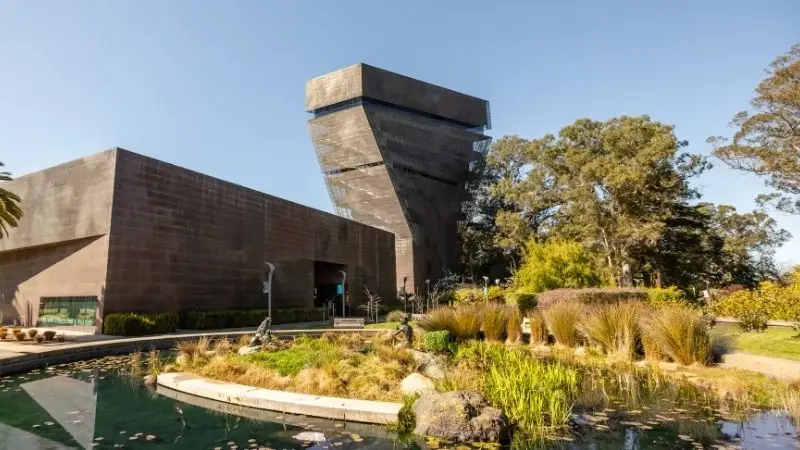 A modern, angular building with a towering structure, seemingly made of dark brick, stands against a clear blue sky. In the foreground, there is a serene pond surrounded by greenery, including tall grasses and aquatic plants. Trees are visible in the background—perfect for exploring Five Ways to Celebrate July 4th in the Bay Area.