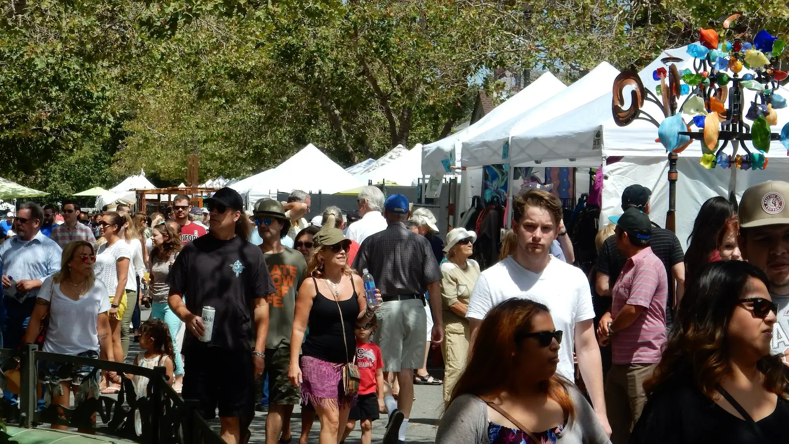 A bustling outdoor fair, one of the best things to do in the South Bay this August, features many people walking past white tents. The crowd includes individuals of various ages, some wearing sunglasses and hats. Trees with green leaves provide shade, and colorful decorations are visible on the right.