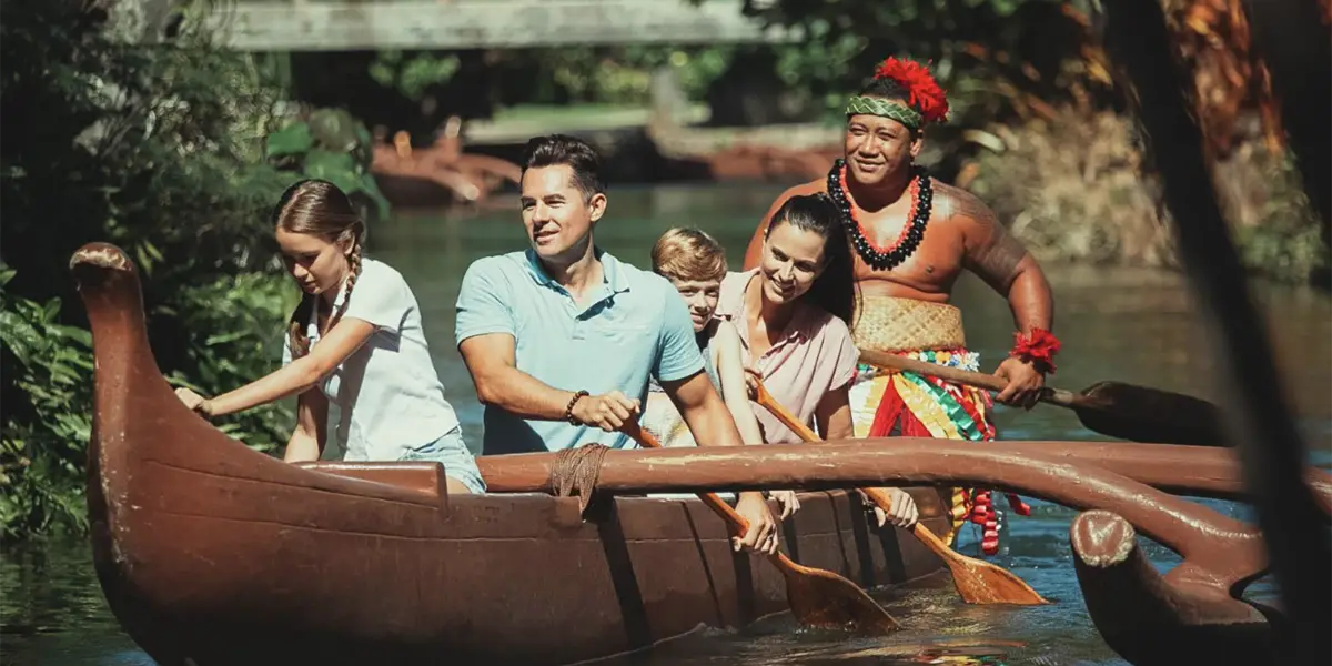 A diverse family of four and a guide paddle a traditional wooden canoe on a calm river. The guide, in festive Polynesian attire with a red headdress and lei, leads the group through lush greenery near a rustic bridge—one of the top family activities Oahu offers.