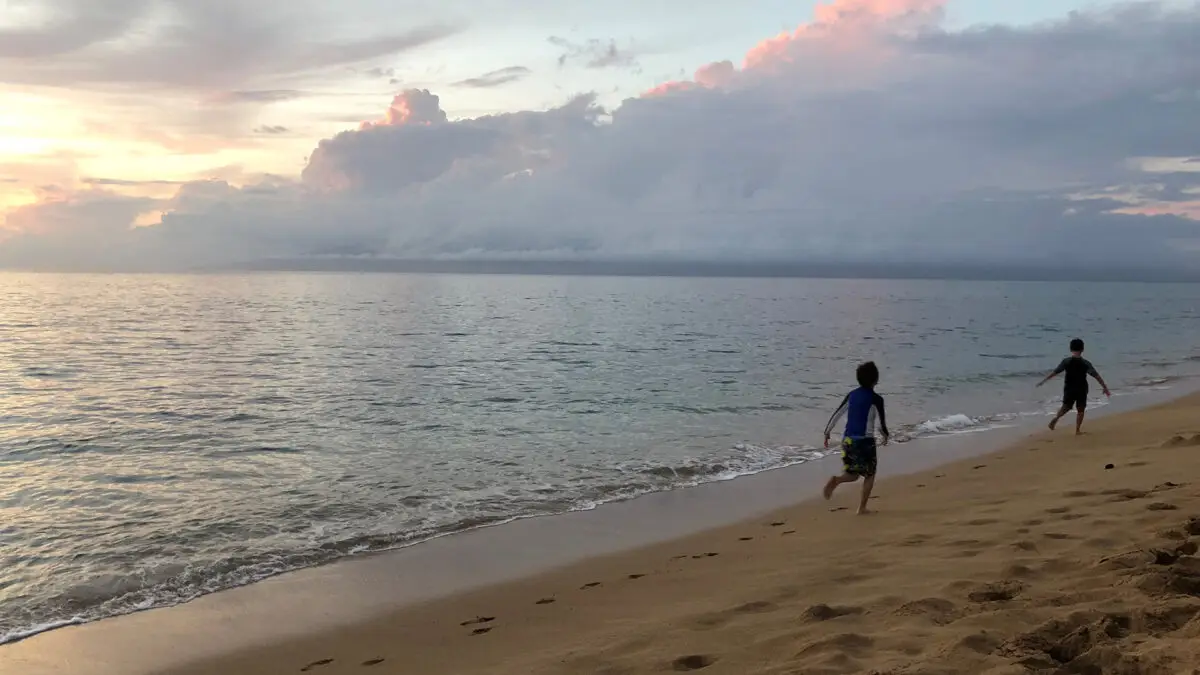 Two children run along a sandy beach at sunset. The ocean is calm with gentle waves lapping the shore. The sky is partly cloudy with hues of pink and orange from the fading sunlight, typical of family beaches in Maui. Footprints are visible in the sand behind the children.