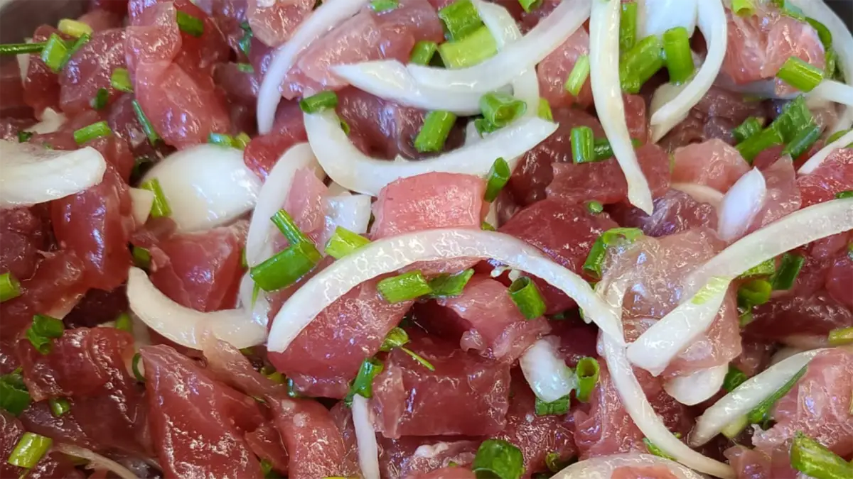 Close-up of a dish featuring raw pieces of tuna marinated with thin slices of onion and garnished with chopped green onions. The vibrant colors and fresh ingredients suggest a dish like poke or ceviche, reminiscent of the best poke in central Kauai.