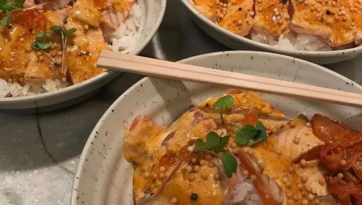 Three bowls of rice topped with slices of seared salmon, garnished with microgreens, roe, and a creamy orange sauce. Each bowl presents a visually appealing and appetizing dish reminiscent of the best poke in Waikiki, Oahu, with a pair of chopsticks resting across one of the bowls.