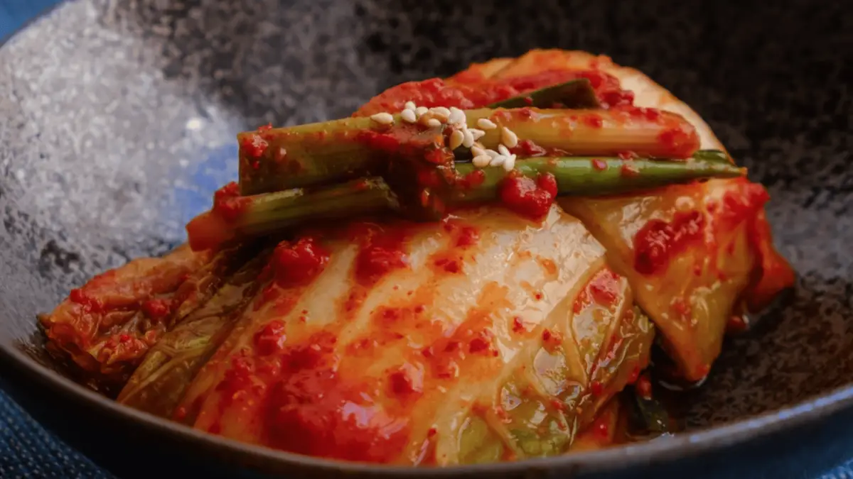 A dish of kimchi presented in a dark bowl. The kimchi is generously coated with spicy red chili paste and garnished with sesame seeds and green onions. The vibrant colors and textures highlight the traditional Korean fermented cabbage dish, showcasing the international influences that shape global cuisine.